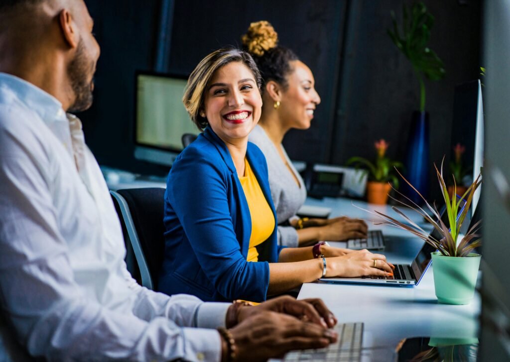 Three people, two women and one man, are seated at a desk, working on computers. The woman in the middle smiles.